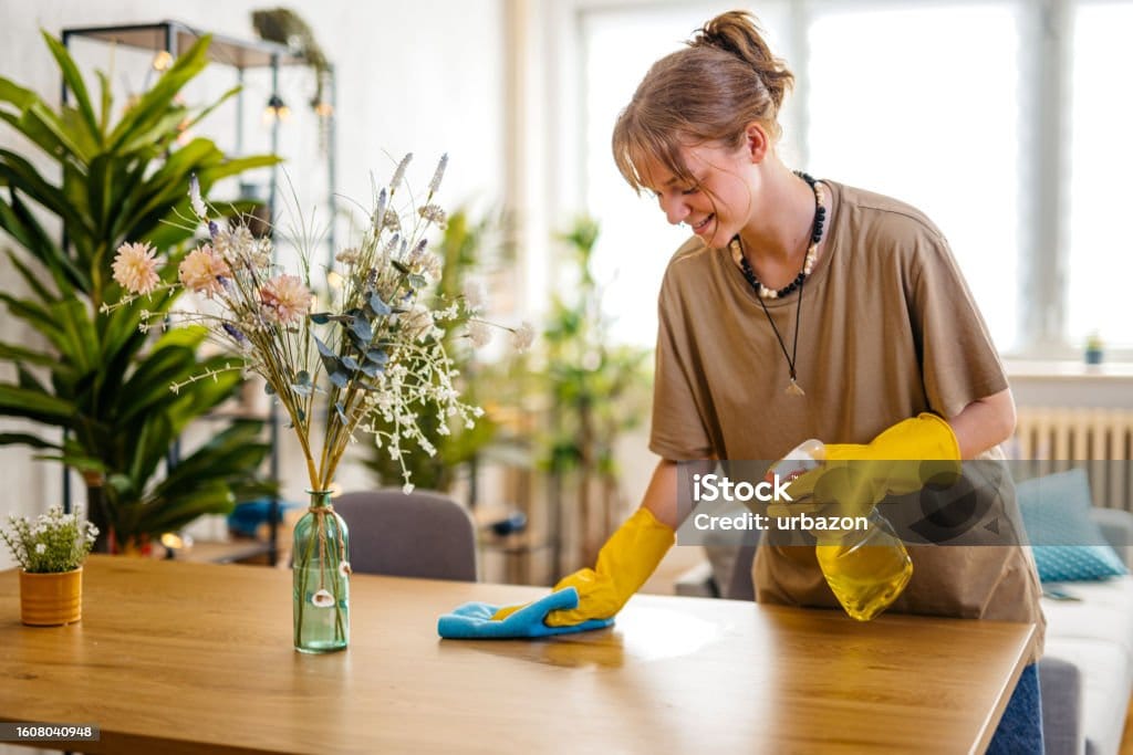 Young smiling woman wiping the dining table surface using a cloth and a spray bottle.